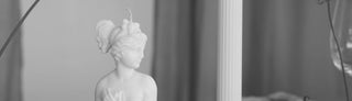 Primo piano della candela Lady Afrodite Ivory accanto a una candela a colonna scanalata, con un calice sullo sfondo, foto in bianco e nero