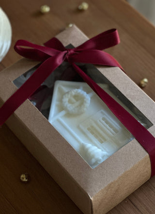 Candela a forma di casetta natalizia visibile nella scatola con finestra trasparente e fiocco bordeaux.