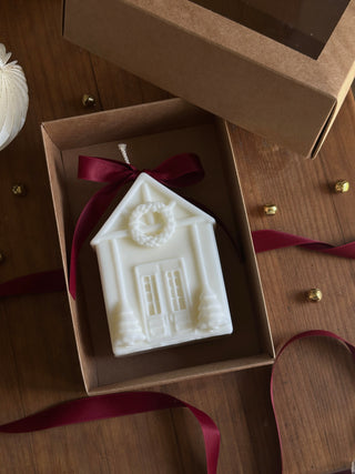 Candela a forma di casetta natalizia in una scatola regalo aperta, con fiocco bordeaux.