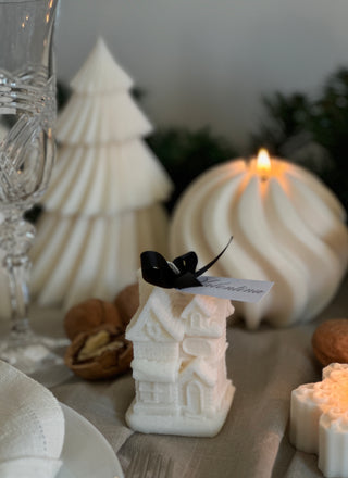 Candela a forma di casetta natalizia su una tavola apparecchiata, accanto ad altre decorazioni e candele natalizie.