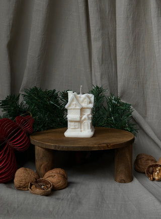 Candela a forma di casetta natalizia su un piccolo tavolino di legno, con decorazioni di Natale sullo sfondo.