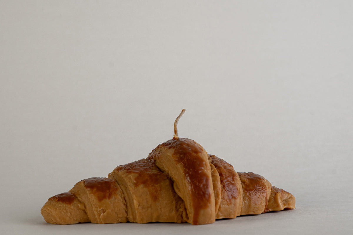 Candela Croissant a forma di cornetto dorato con stoppino, vista laterale su fondo grigio chiaro