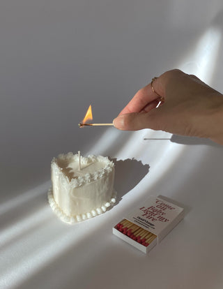 Una mano con un anello dorato accende la candela a forma di cuore accanto a una scatola di fiammiferi.