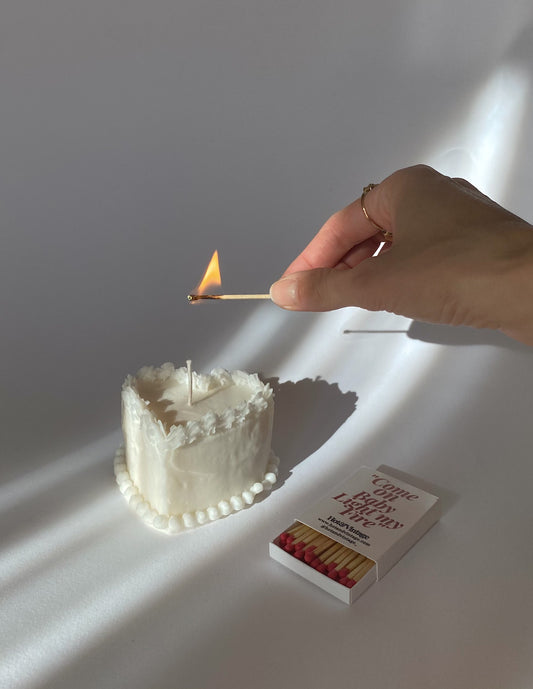 Una mano con un anello dorato accende la candela a forma di cuore accanto a una scatola di fiammiferi.