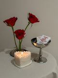 Candela a forma di cuore accesa accanto a rose rosse e a una coppetta con fiammiferi.