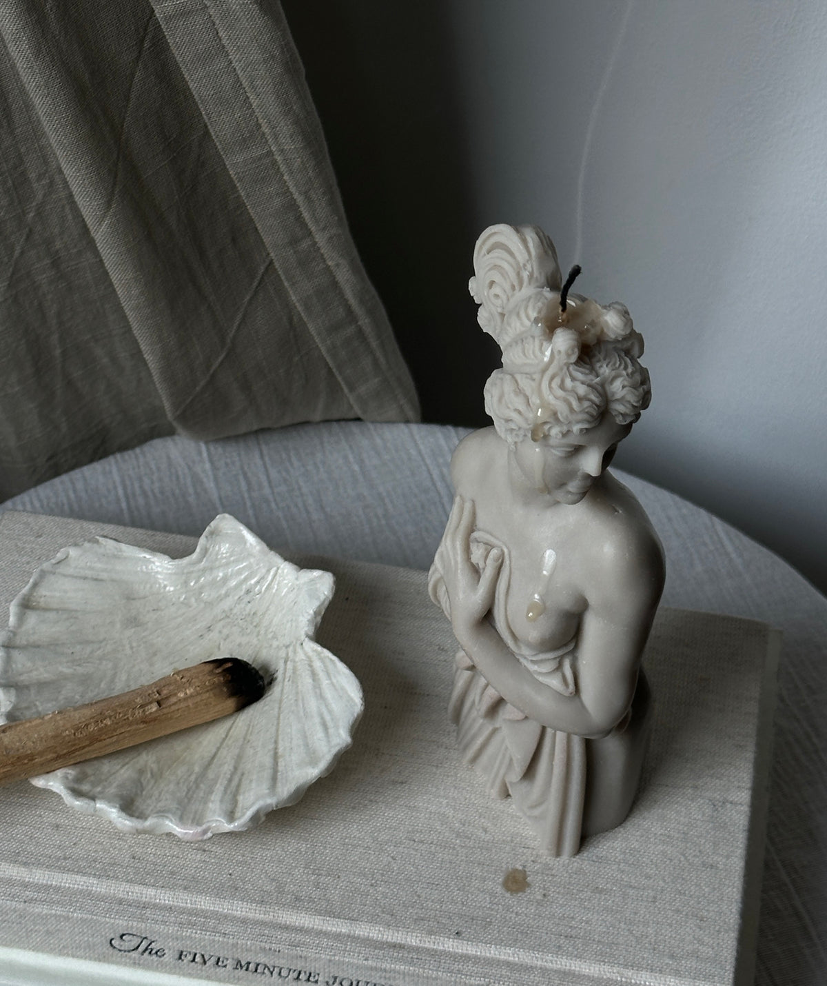 Candela Lady Aphrodite color grigio pietra su un libro accanto a una conchiglia.