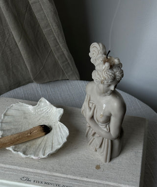 Candela Lady Aphrodite color grigio pietra su un libro accanto a una conchiglia.