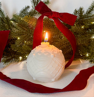 Candela a forma di pallina di Natale accesa, con foglie verdi e fiocco rosso sullo sfondo.