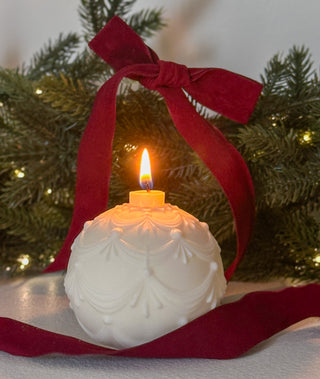 Candela a forma di pallina di Natale accesa, con foglie verdi e fiocco rosso sullo sfondo.