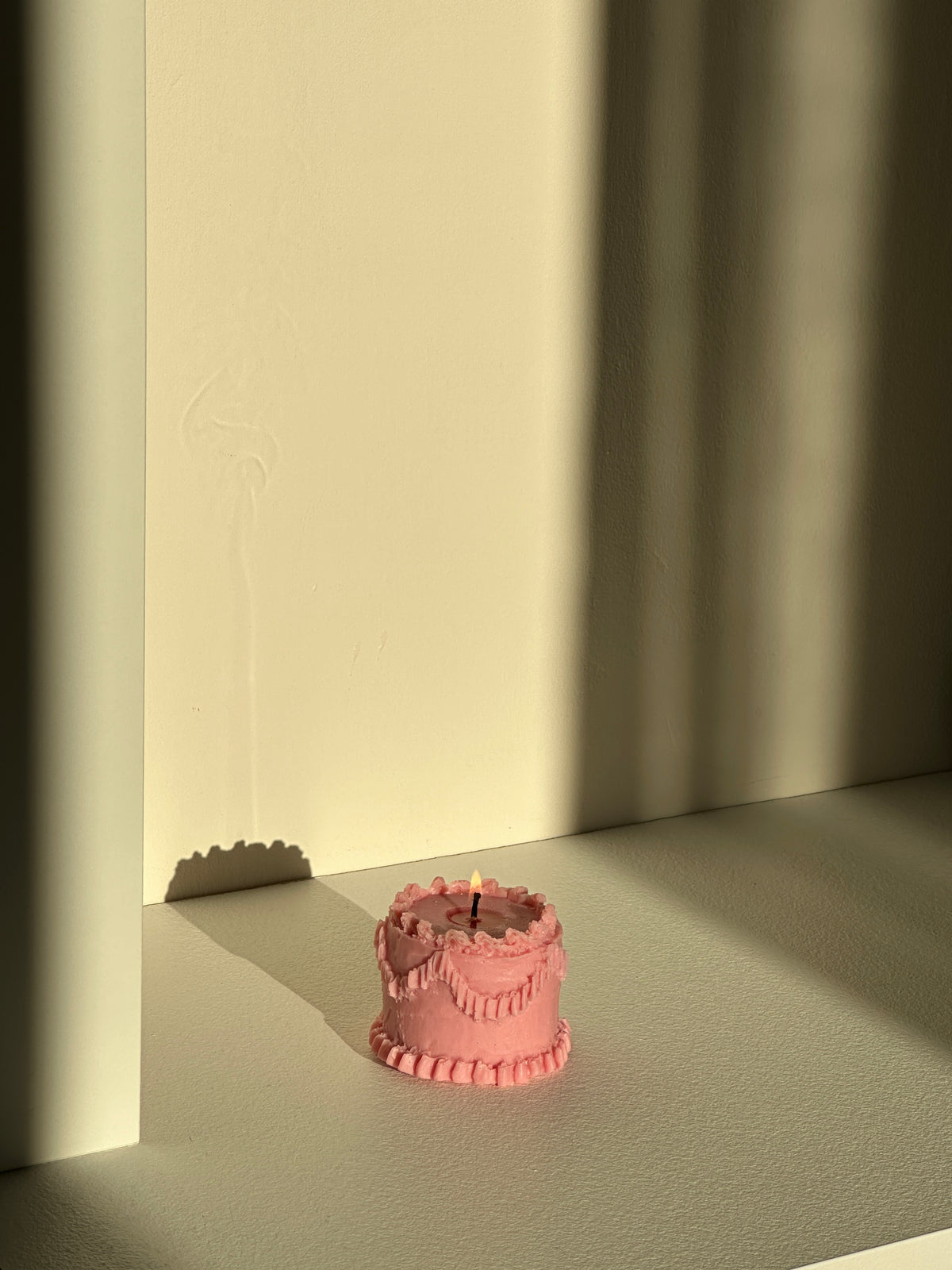 Candela rosa a forma di torta accesa davanti a una parete illuminata dalla luce calda del tramonto.