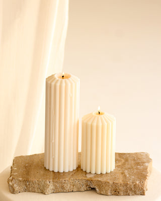 Due candele a colonna color avorio, una piccola e una grande, appoggiate su una lastra di marmo con una tenda chiara sullo sfondo.