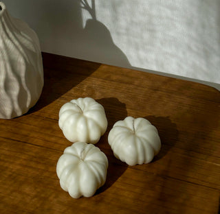Candele di Halloween a forma di zucca color avorio e terracotta su un tavolino di legno, con una tazza e pinoli sullo sfondo.