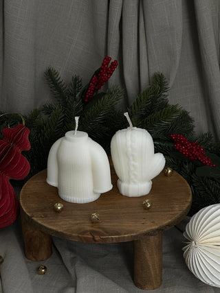 Candele a forma di maglione e muffola su un tavolo in legno, con campanelli e rami verdi su fondo grigio.