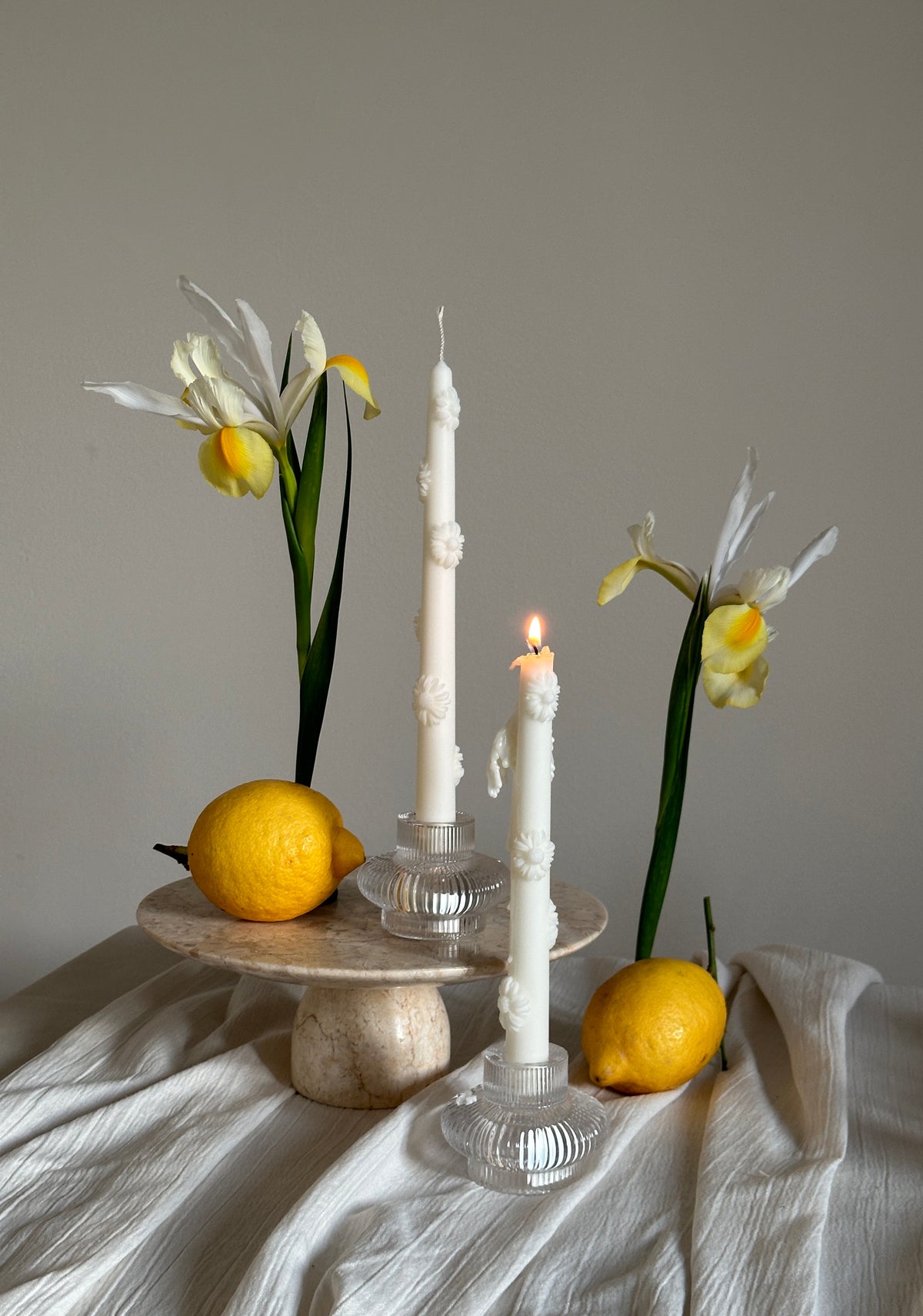 Due candele alte a forma di margherita accanto a limoni e fiori, su un tavolo di marmo.