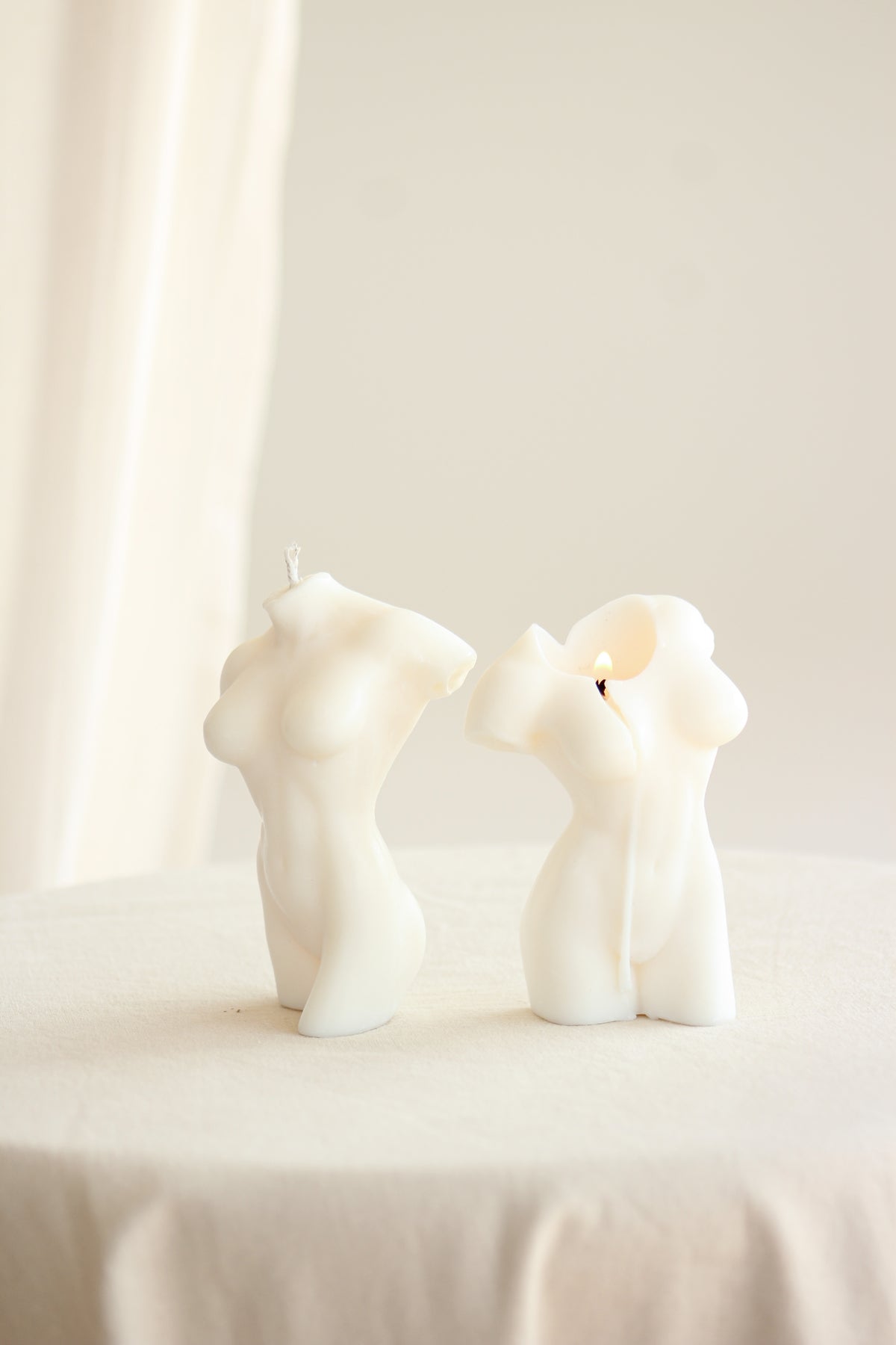 Due candele Muse color avorio a forma di busto femminile, una accesa e una spenta, su una tovaglia bianca.