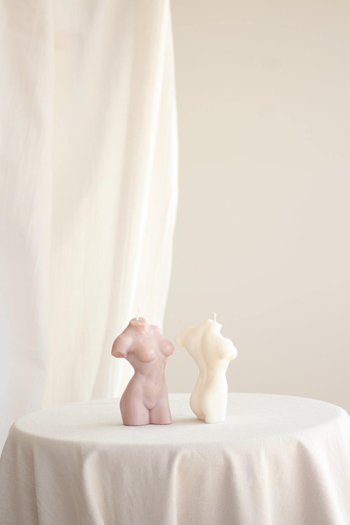 Due candele Muse a forma di busto femminile, una Dusty Pink e una avorio, affiancate su una tovaglia bianca.