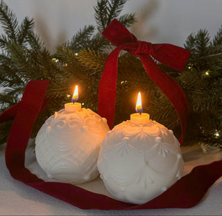 Coppia di candele a forma di pallina di Natale accese, con foglie verdi e fiocco rosso sullo sfondo.