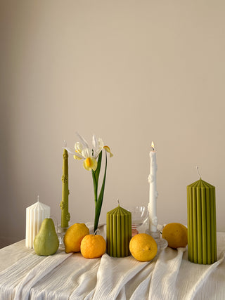 Composizione di candele a forma di frutta nei toni del verde, giallo e bianco, con limoni e pere su un tavolo chiaro.