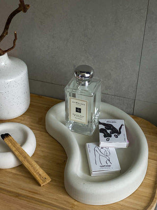Due scatole di fiammiferi illustrate Astrid Babayan su un vassoio bianco, accanto a un profumo, su un tavolo di legno.