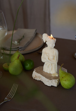 Candela Lady Afrodite Ivory accesa su una base in pietra, su una tavola apparecchiata con pere, lime, piatto e tovagliolo