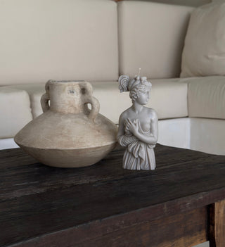 Candela Lady Afrodite Stone Grey, busto in cera grigio tortora accanto a un vaso in terracotta su un tavolo di legno scuro