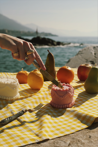 Mano che taglia con un coltello una candela a torta rosa, accanto a una candela a torta bianca, frutta e un telo a quadretti gialli sul mare