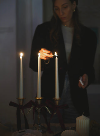 Donna che accende con un fiammifero tre candele lunghe su un candelabro con nastri di velluto bordeaux, su una tavola apparecchiata