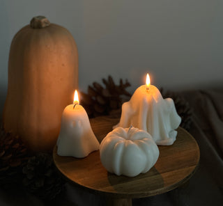Due candele a forma di fantasma accese e una candela a forma di zucca bianca su un piano di legno, con zucche e pigne sullo sfondo