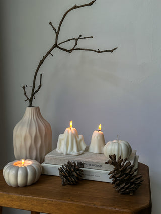 Due candele a forma di fantasma accese e una candela a zucca, tutte Ivory, su dei libri con vaso, ramo secco e pigne