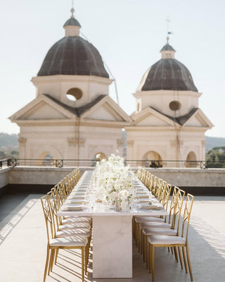 Lunga tavola di marmo per un matrimonio su una terrazza, con fiori bianchi e candele Lady Afrodite al centro, sedie dorate e due cupole di una chiesa sullo sfondo