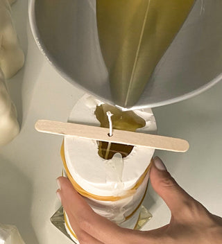 Colata della cera con un imbuto nello stampo bianco tenuto fermo da elastici, con lo stoppino fissato da uno stecchetto di legno