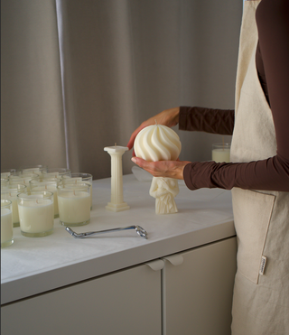 Donna con grembiule di lino che tiene in mano una candela a spirale Ivory nel laboratorio, accanto a una candela a colonna e a vasetti di candele
