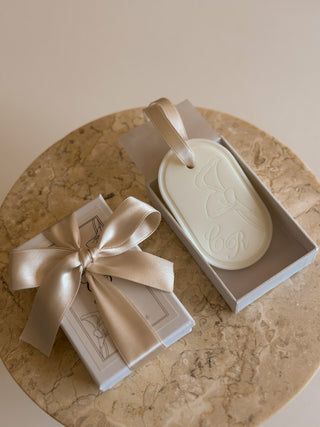 Tavoletta profumata ovale con calla e iniziali incise, per matrimonio, accanto alla scatola chiusa con fiocco di raso su un piano in marmo