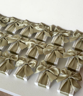 Confezioni regalo per matrimonio, scatole bianche con tavoletta profumata e fiocco di raso verde salvia, disposte in file su un piano bianco