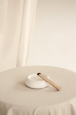 Piattino rotondo in Jesmonite con un piccolo accessorio in legno per bruciare incenso, su una tovaglia chiara.
