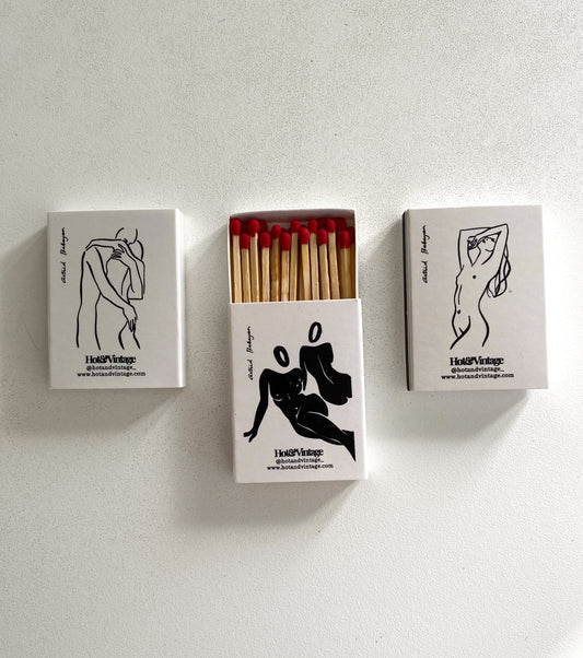 Tre scatole di fiammiferi illustrate da Astrid Babayan, con quella centrale aperta e i fiammiferi visibili.