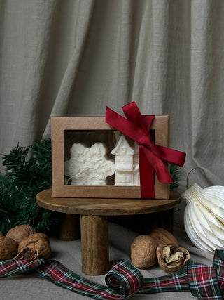 Candela a forma di fiocco di neve e casetta natalizia confezionate in una scatola regalo chiusa, con decorazioni e schiaccianoci sullo sfondo.