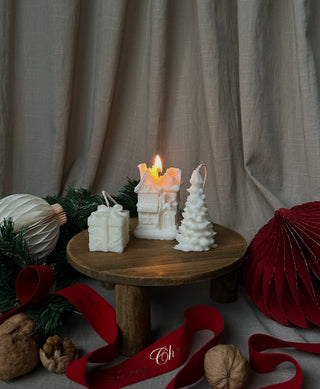 Set di tre mini candele natalizie senza confezione, con la casetta accesa al centro, tra un pacchetto regalo e un alberello.