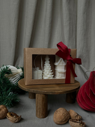 Set di tre mini candele natalizie, con pacchetto regalo, casetta e albero, confezionato con un fiocco rosso e circondato da decorazioni natalizie.